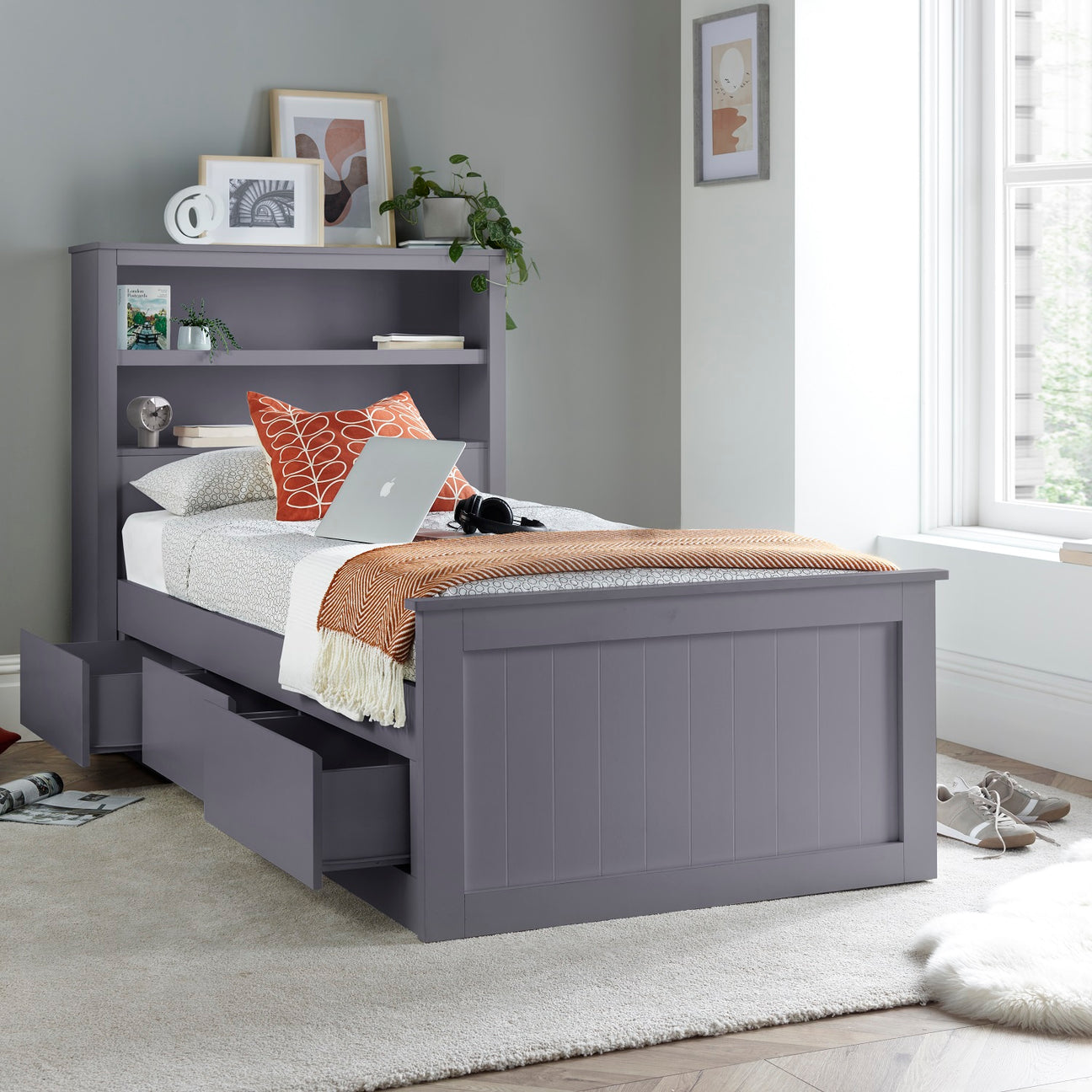 A grey single bed frame with built-in bookshelves and under-bed storage drawers, styled in a modern bedroom setting with a white rug, window, and decorative pillows.