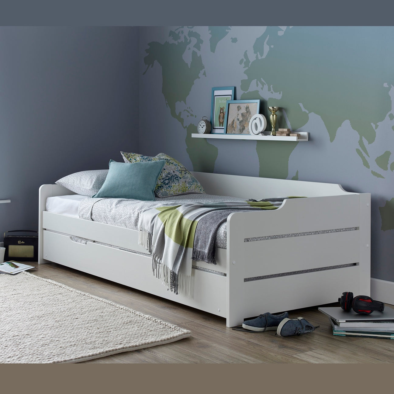 A white wooden daybed frame with under-bed trundle, styled with bedding and pillows in a bedroom setting with a world map wall mural.