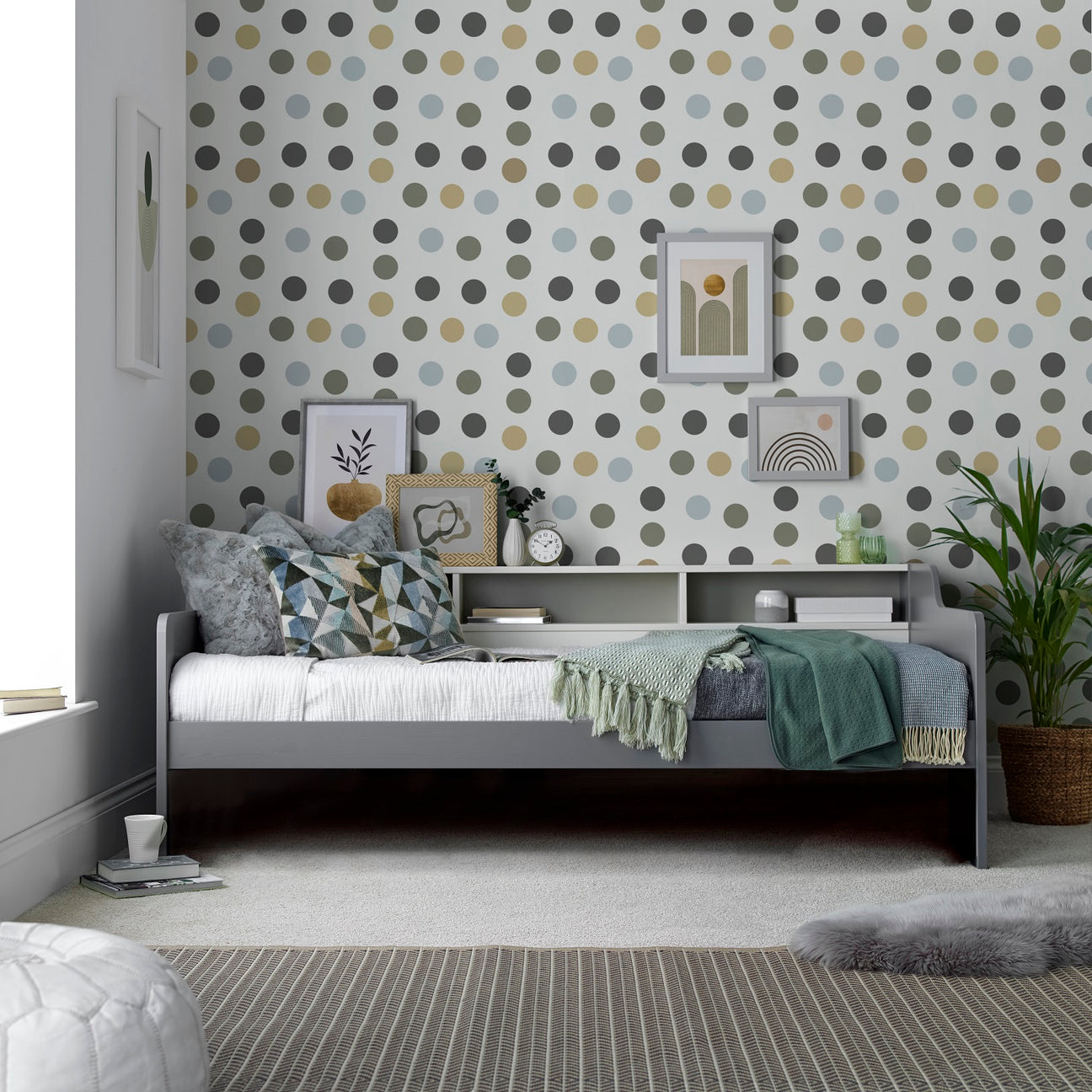 A styled bedroom interior featuring a two tone grey/white daybed with patterned pillows and a green throw blanket, set against a wall covered in white wallpaper with a repeating polka dot pattern in muted grey, olive green, and beige. The room includes framed wall art, a small shelf, a potted plant, and a striped rug.