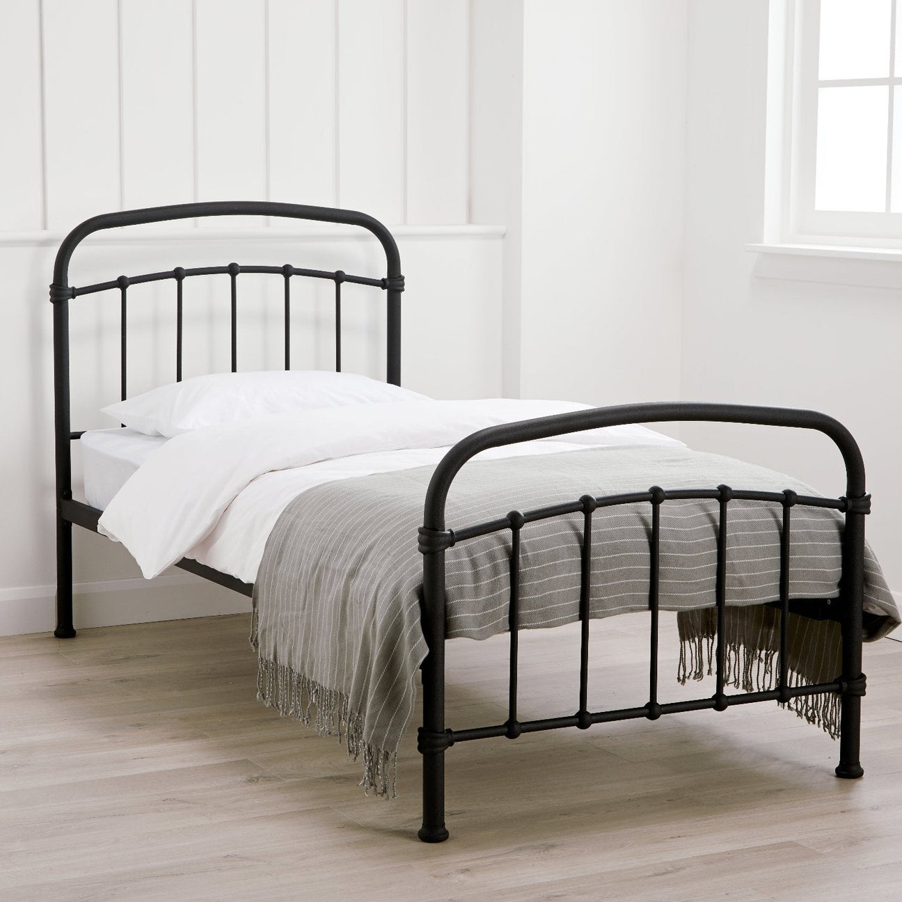 Halston Metal Bed Frame - Black metal bed frame with rounded headboard and footbaord and vertical bars, in a bright modern bedroom, positioned at an angle- Beds4Us